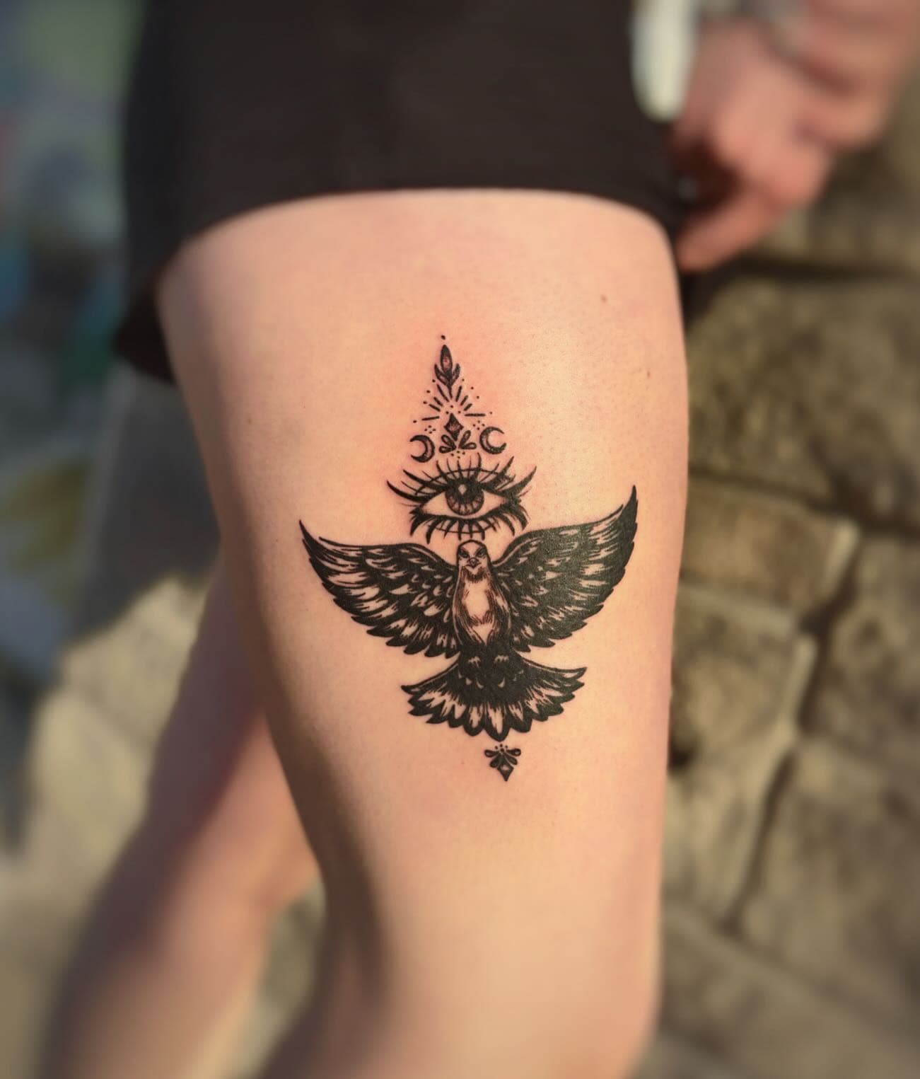 Black ink tattoo of an owl with an all-seeing eye and moon phases on a person's thigh