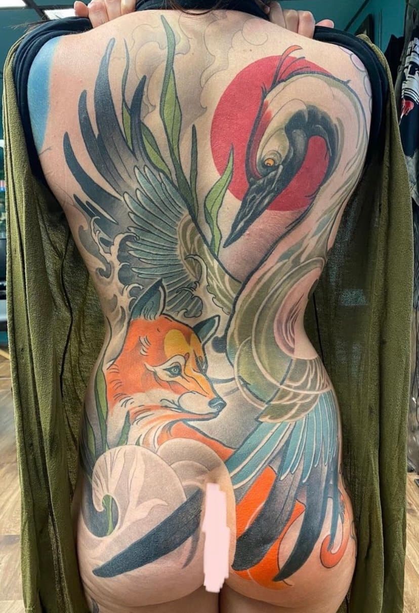Colorful tattoo of a crane with a red sun, a fox, and flowing waves and leaves on a person's back and lower body.