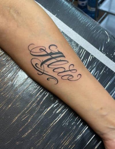 Forearm tattoo of a personal name in flowing black cursive script lettering