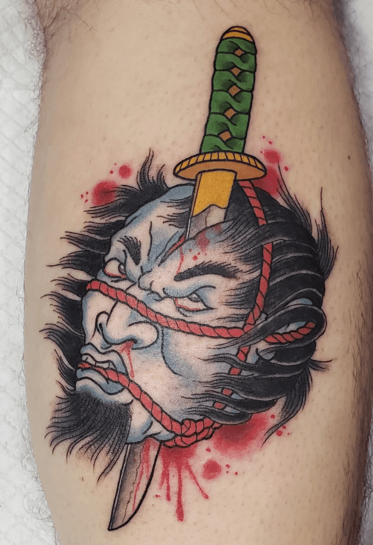 Tattoo of a person's face split into two halves with a knife through the middle, surrounded by red splatters, with a green and yellow dagger stabbed through the