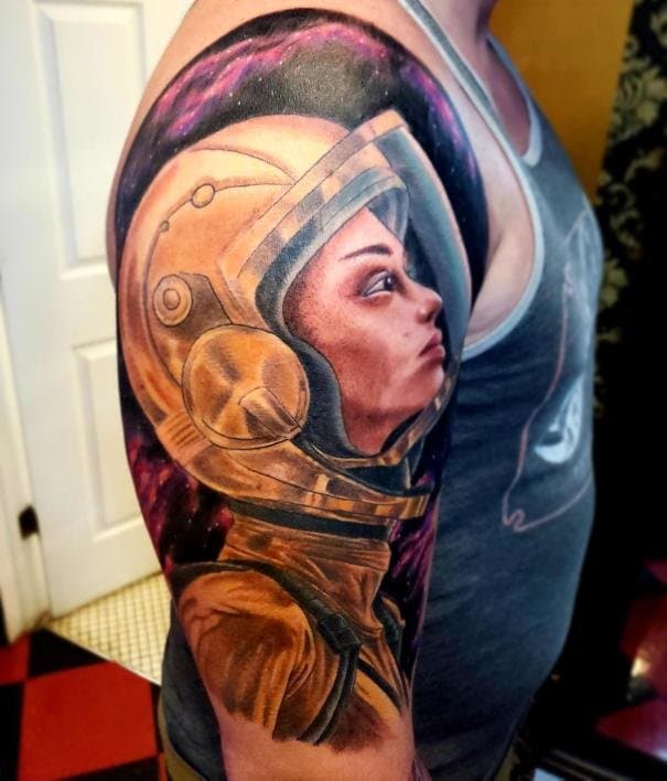 Tattoo of a woman's face in an astronaut helmet on a person's upper arm, with a space background.