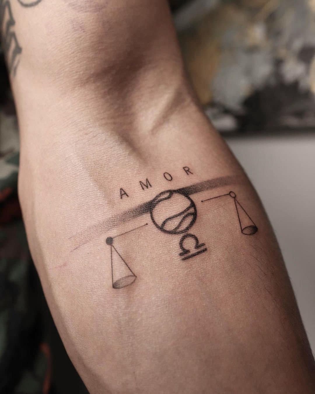 Fine line inner-arm tattoo of balanced scales with the word AMOR