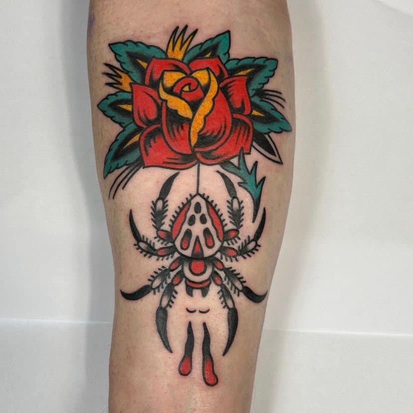 Tattoo of a red and yellow rose above a spider with a skull on its back, in traditional style with bold black outlines and red, yellow, green, and gray colors.