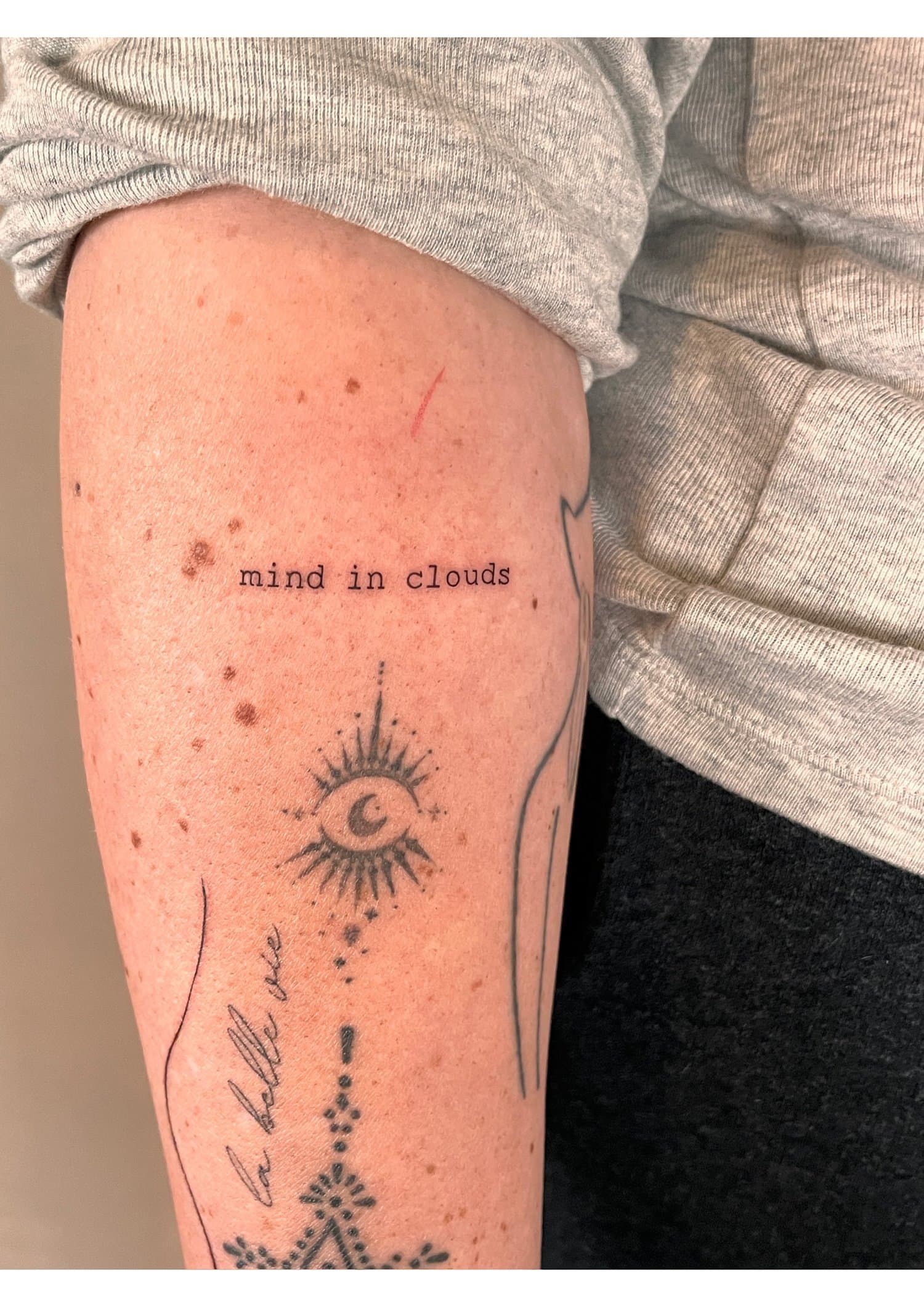 Close-up of a person's upper arm with tattoos, including a sun with a moon inside and text reading 'mind in clouds', wearing a gray long-sleeve shirt.