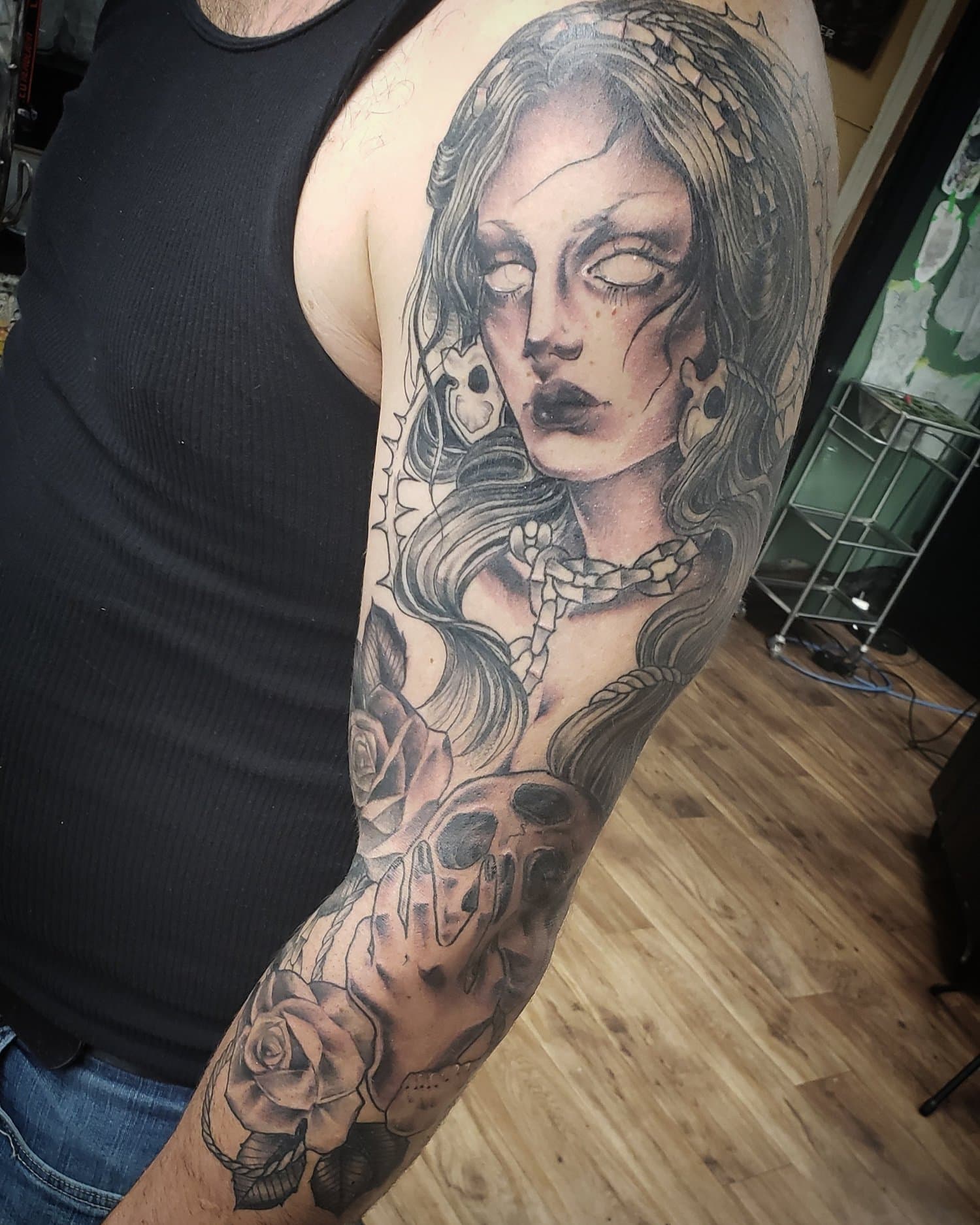 Tattoo of a woman with long hair, blank eyes, and dark makeup, surrounded by roses, a snake, and a skull on a person's upper arm.
