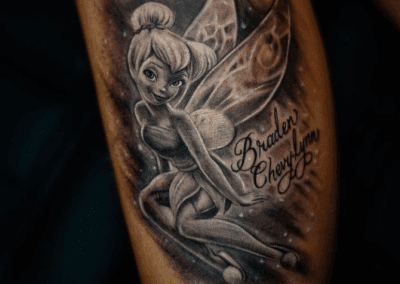 Tinkerbell tattoo in black and grey ink features the fairy with wings, seated next to stylized script reading "Braden Chevylynn".