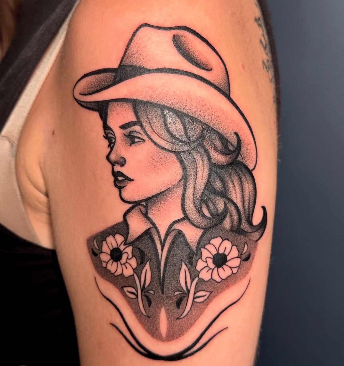 Black-and-grey illustrative cowgirl portrait tattoo on an upper arm, attributed to Leah on her Jade and Dagger profile page