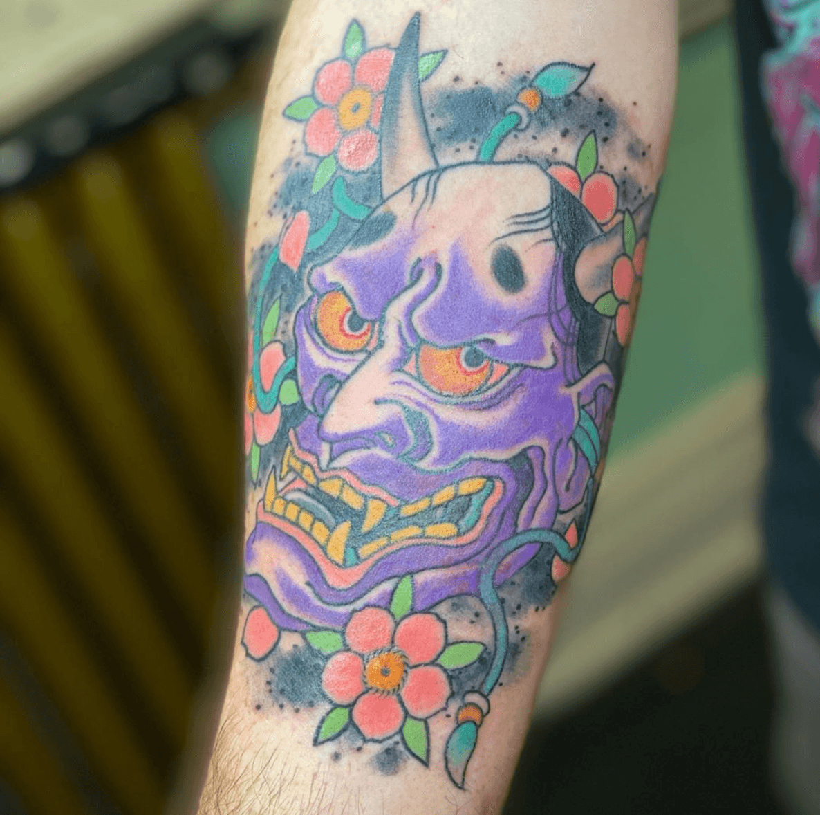 Colour purple oni-mask tattoo framed by red flowers and dark brush-like shading