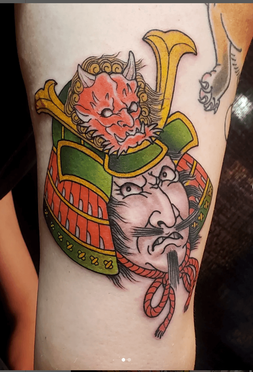 Tattoo of a fierce samurai warrior with a demon mask on his helmet, wearing traditional armor and with a stern facial expression.