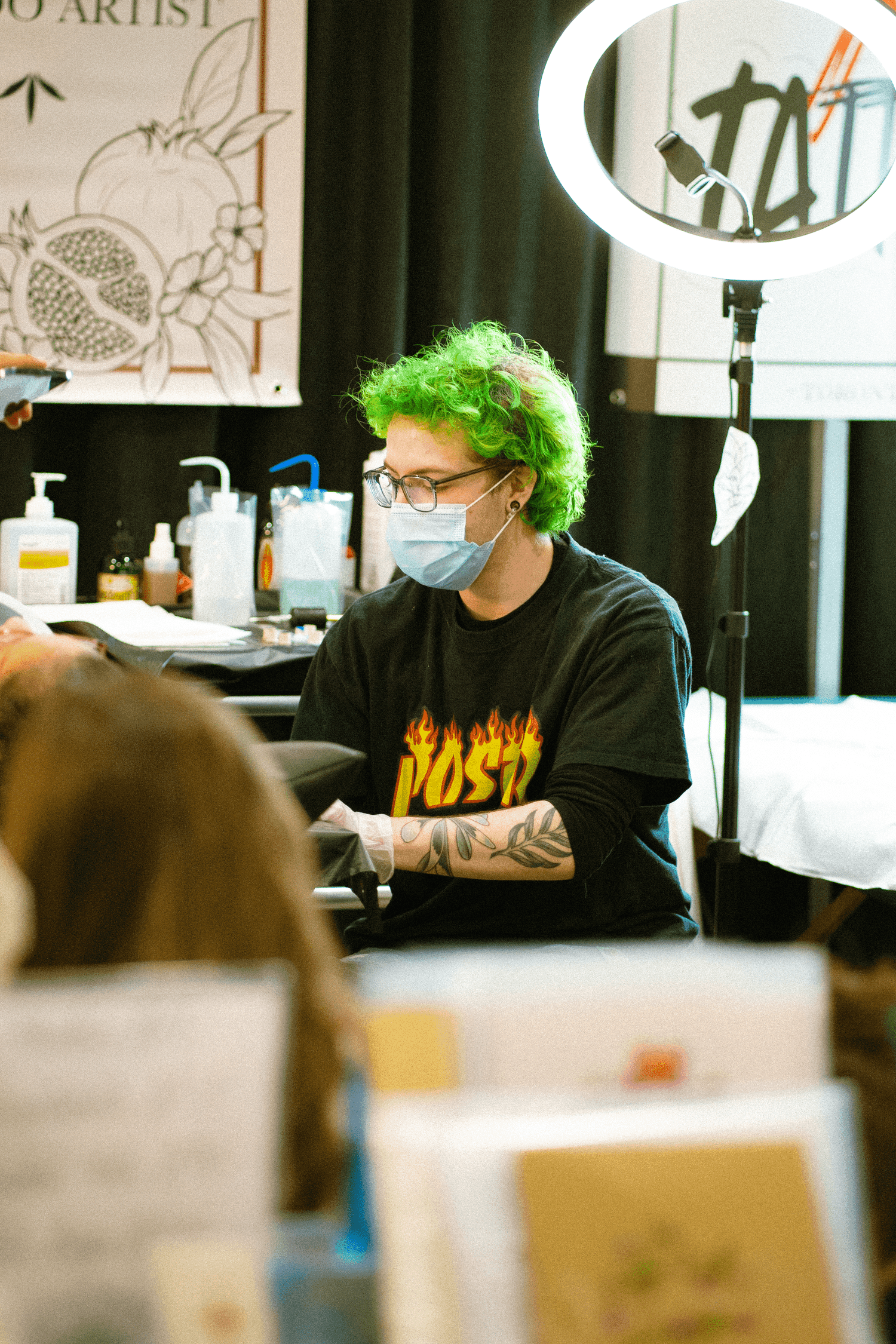 A person with bright green hair, glasses, and a face mask tattooed arm, working at a table with tattoo supplies and a ring light.