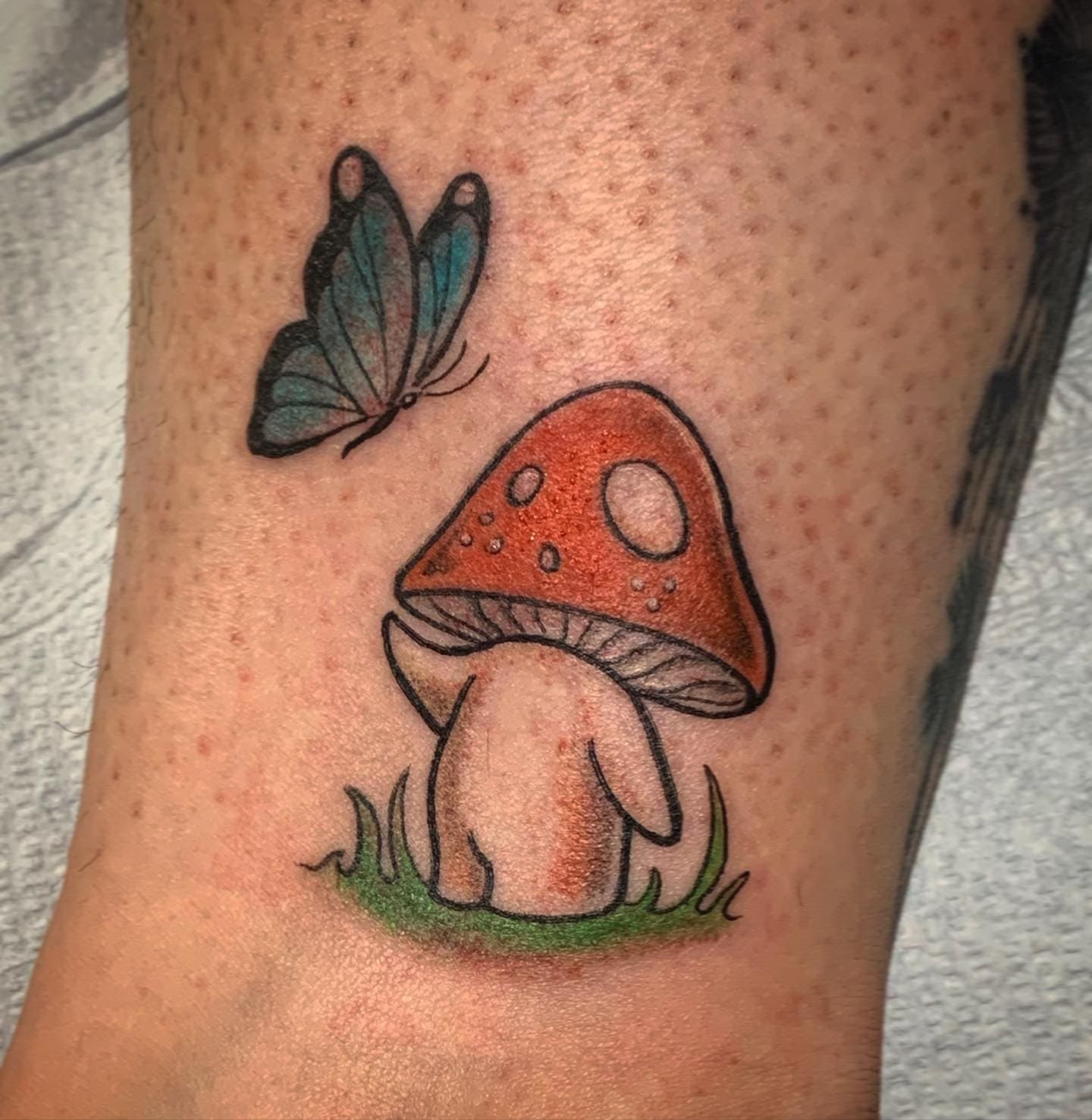 Tattoo of a cartoon mushroom with a red cap, and a blue butterfly flying nearby on skin with visible freckles.