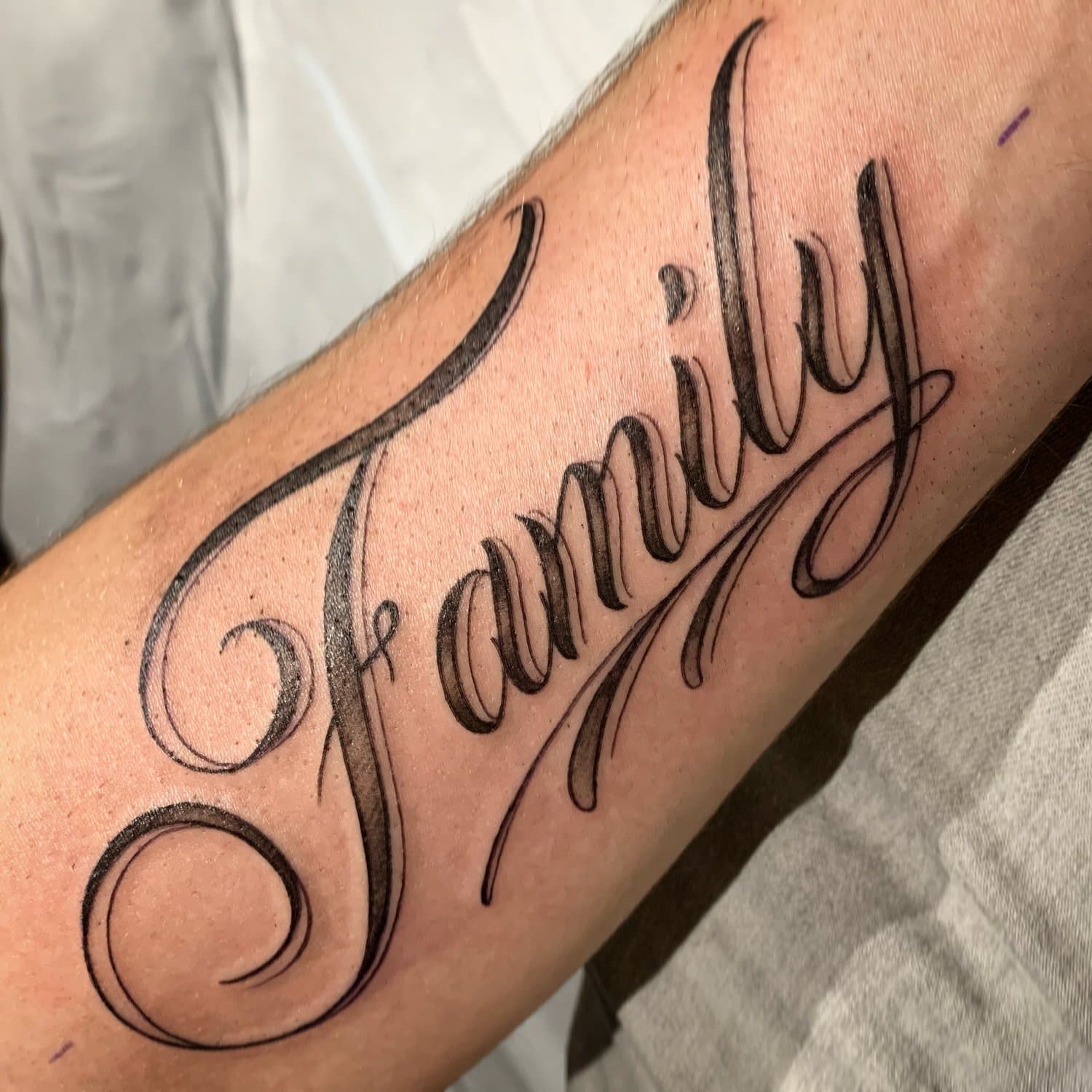 Tattoo of the word "Family" in elaborate cursive script on person's forearm.