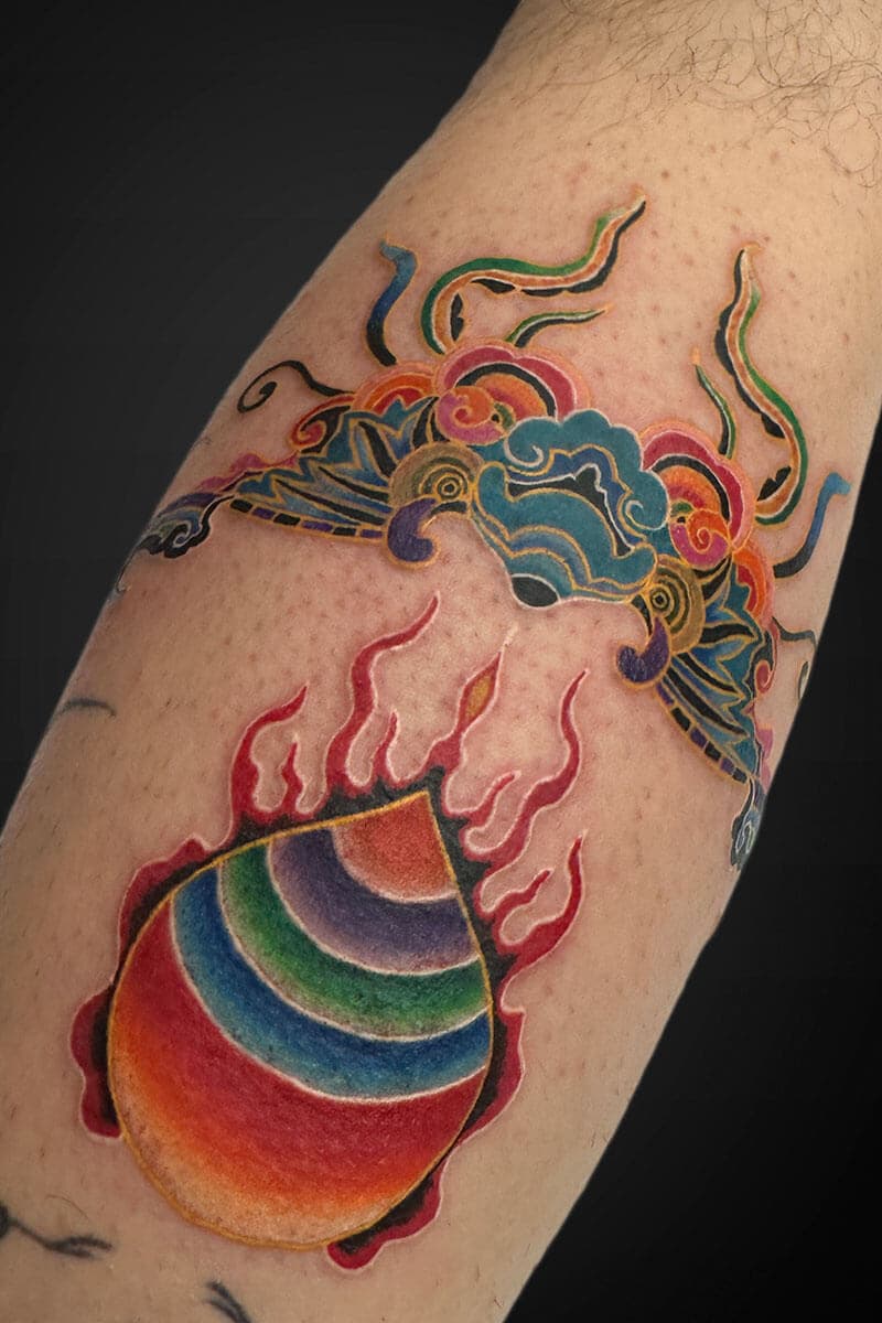 Colorful rainbow orb with red flames surrounding it, tattooed on a forearm, with decorative blue and green cloud motifs above.