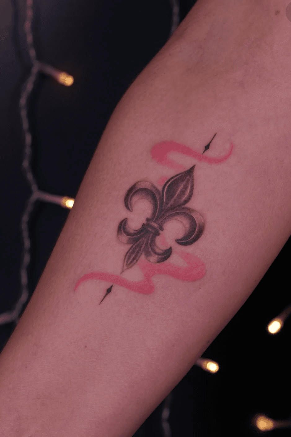 Small black-and-grey fleur-de-lis tattoo with red accent lines on a forearm, attributed to Eunseo on her Jade and Dagger profile page