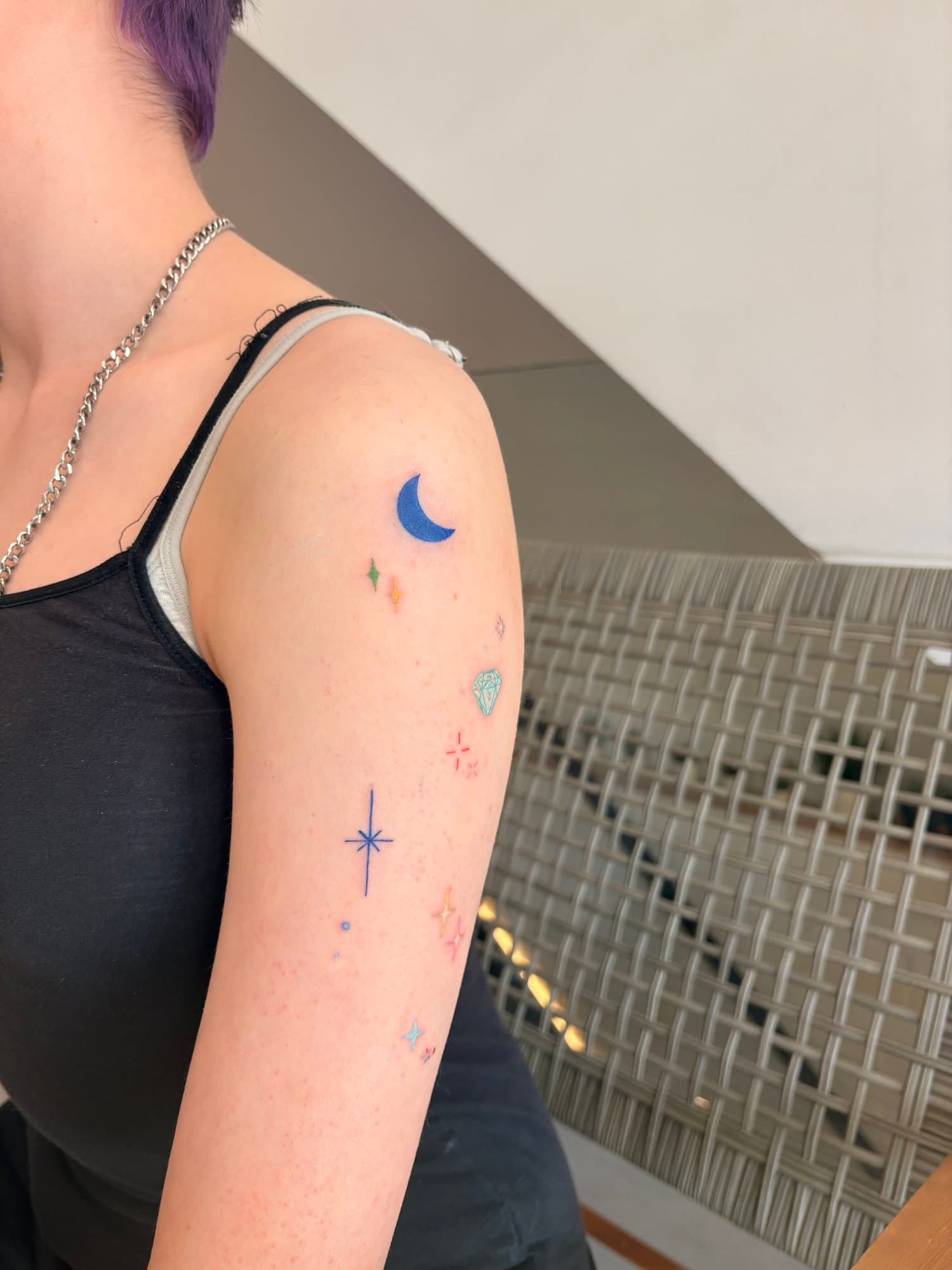 Close-up of a person's upper arm with colorful small tattoo stickers, including a blue crescent moon and various star-shaped designs, with a background of a til