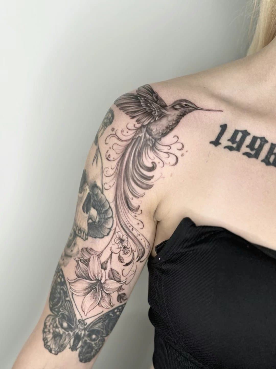 Black-and-grey hummingbird with long ornamental feathers and flowers across the shoulder and upper arm