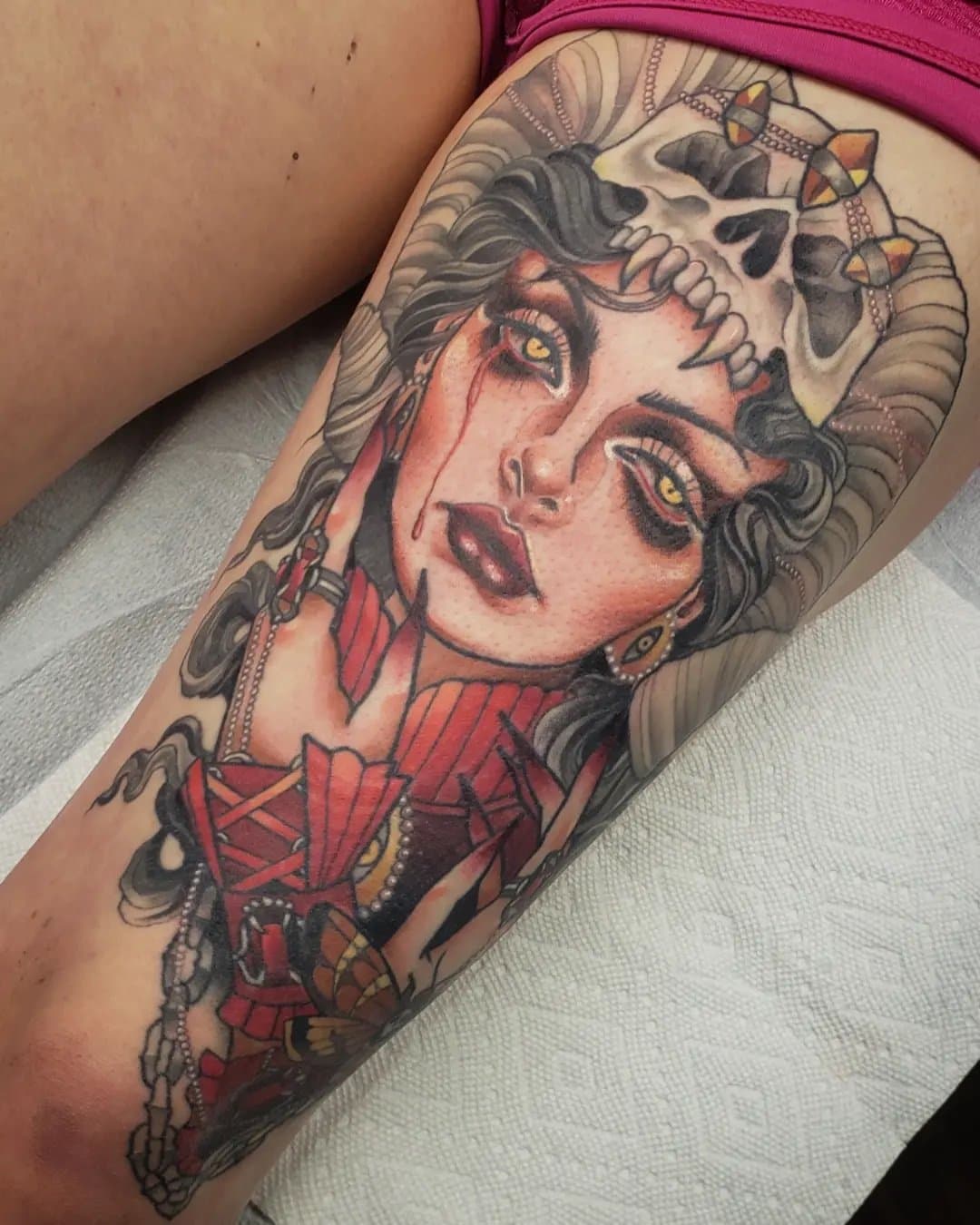 Tattoo of a woman with dark hair, yellow eyes, and blood tears, wearing a skull crown and red clothing, with dark makeup.