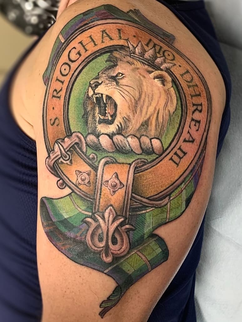 Colour upper-arm tattoo of a Scottish clan crest with a crowned lion's head inside a lettered belt and buckle over a tartan sash