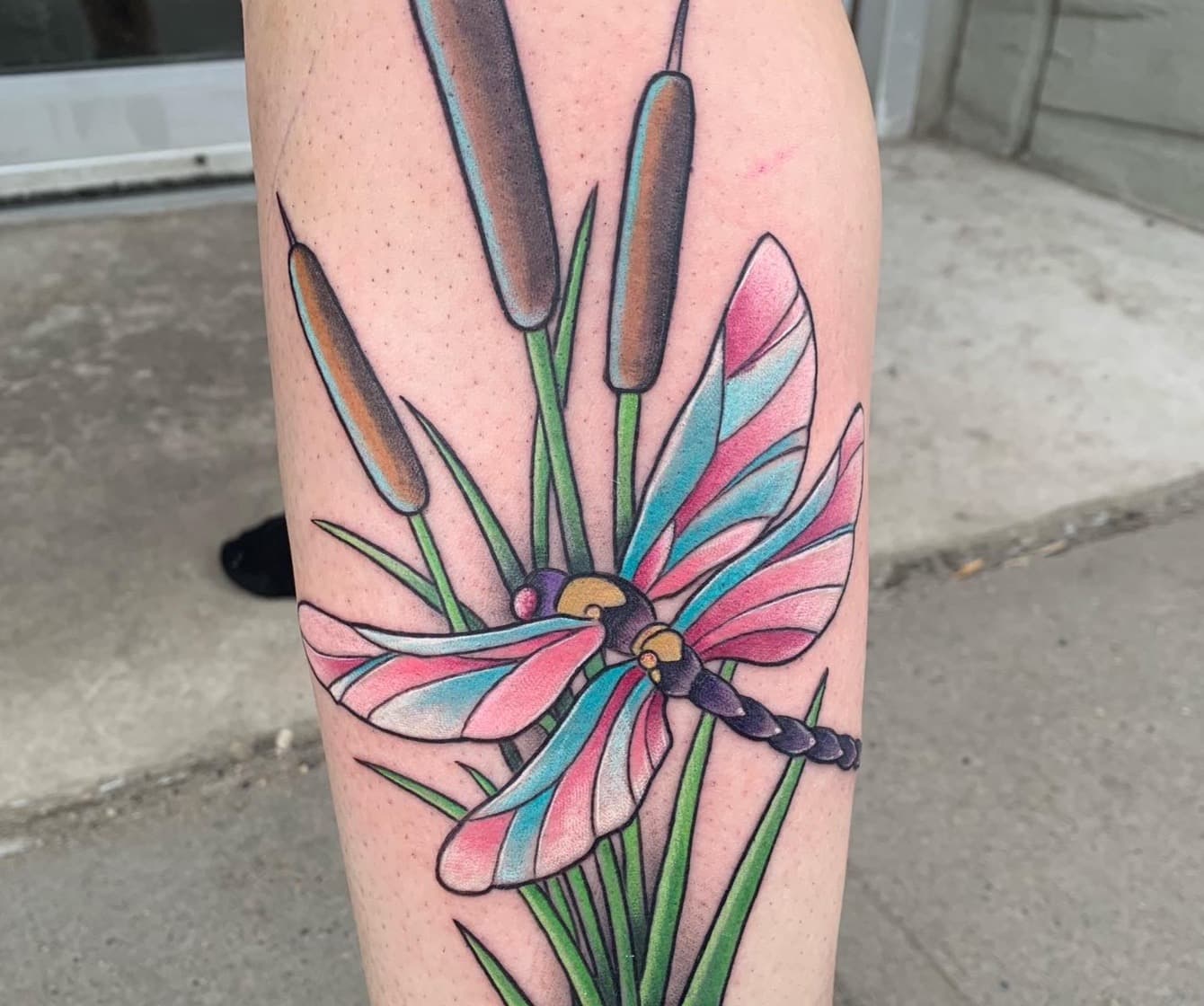 Full-colour tattoo of a pink and blue dragonfly over green cattails on a lower leg