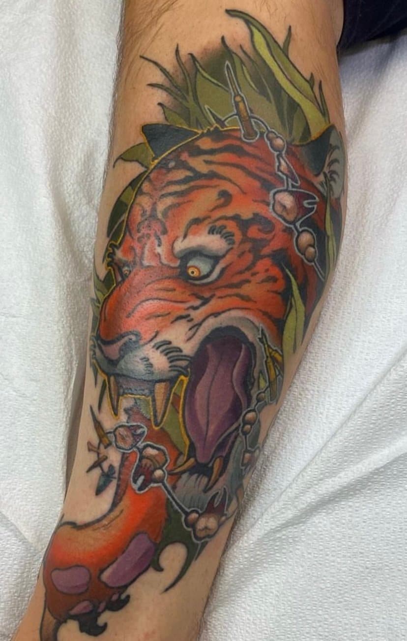 Tattoo of a roaring tiger's head with sharp teeth, set among green leaves and chains.