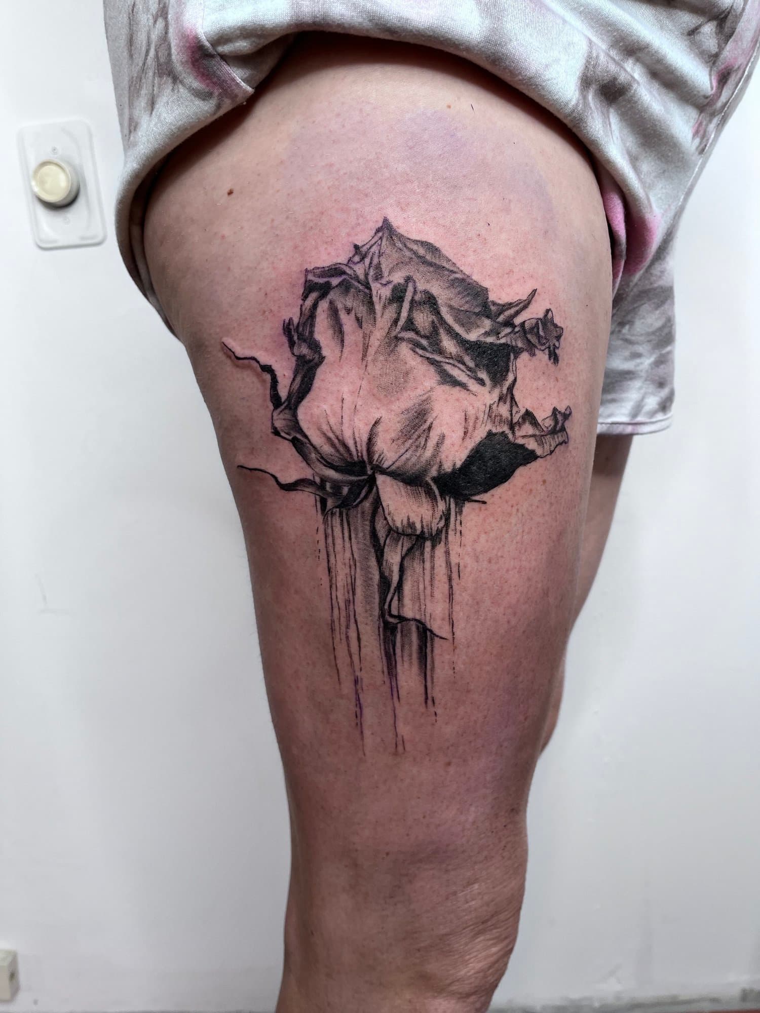 Black ink tattoo of a rose on a person's upper thigh with sketchy, watercolor-style shading and dripping effects.