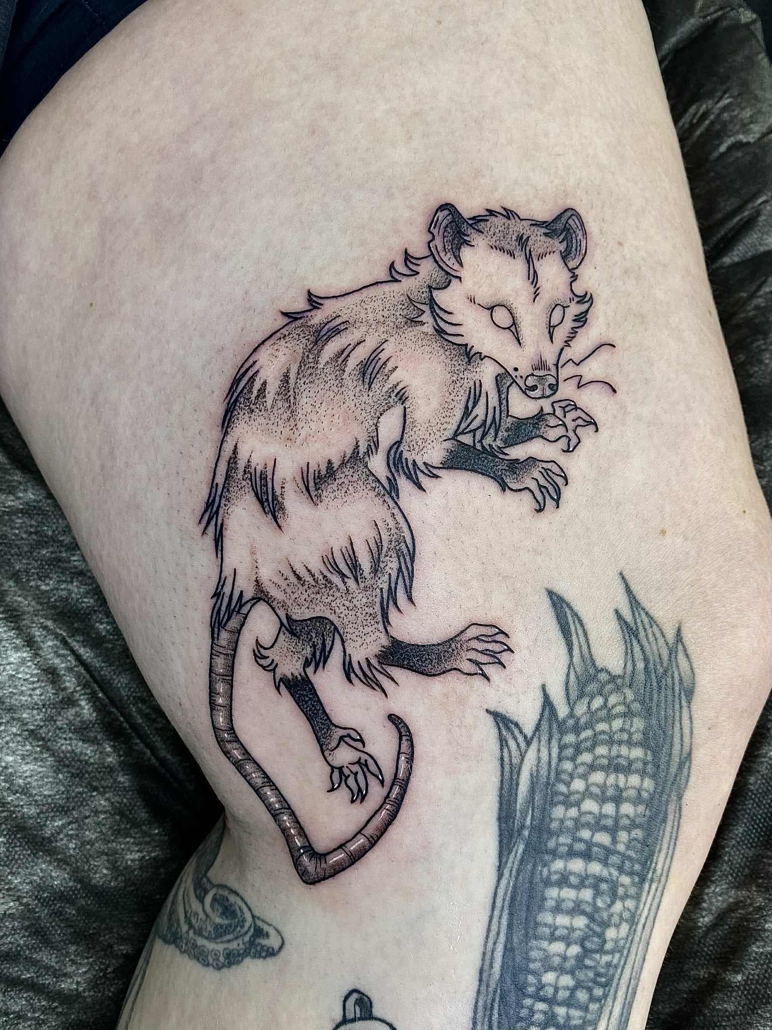 Black-and-grey thigh tattoo of an opossum climbing beside a corn cob