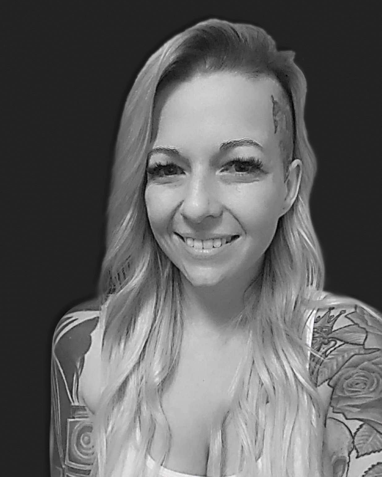 Black and white photo of a woman with long wavy hair, smiling, showing her teeth, and tattoos on her shoulders and arms.
