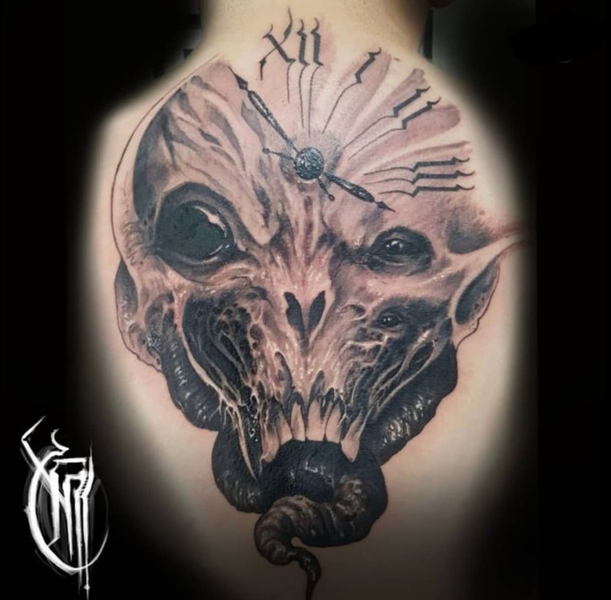 Black and grey horned skull-creature tattoo with a clock face above it, on an inner arm, carrying the artist's monogram watermark