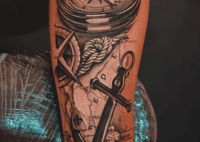 Forearm tattoo shows a realistic compass, nautical rope, anchor, and map with cities like Stockholm and place names in black ink.