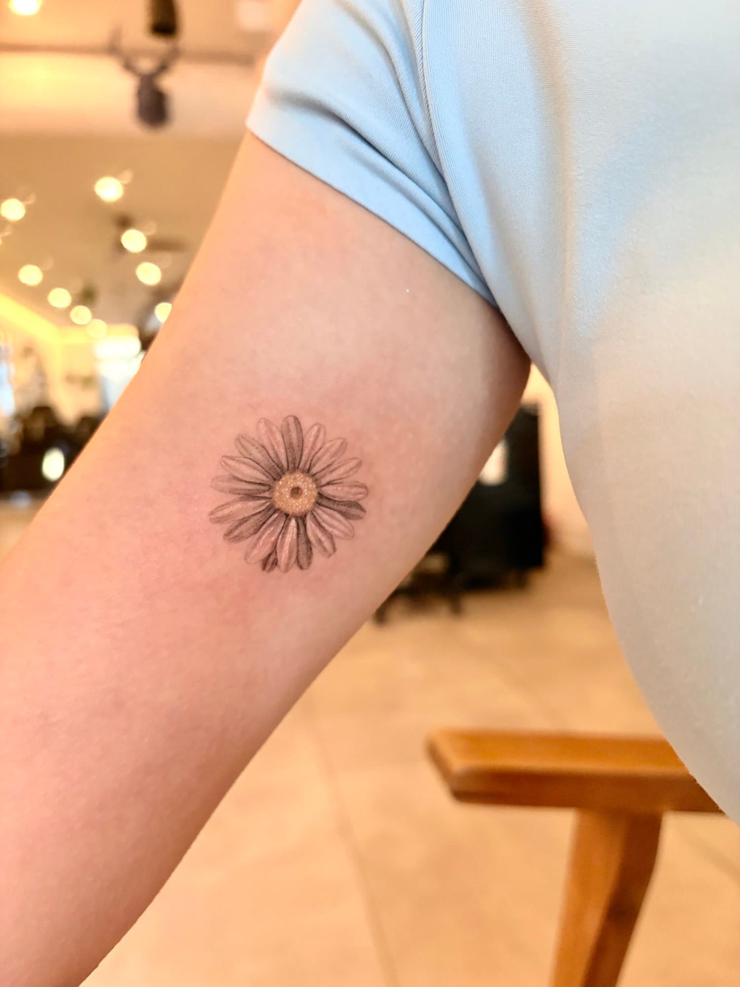 Close-up of a person's upper arm showing a tattoo of a daisy flower on the inner forearm, wearing a light blue short-sleeved shirt, indoor background with warm