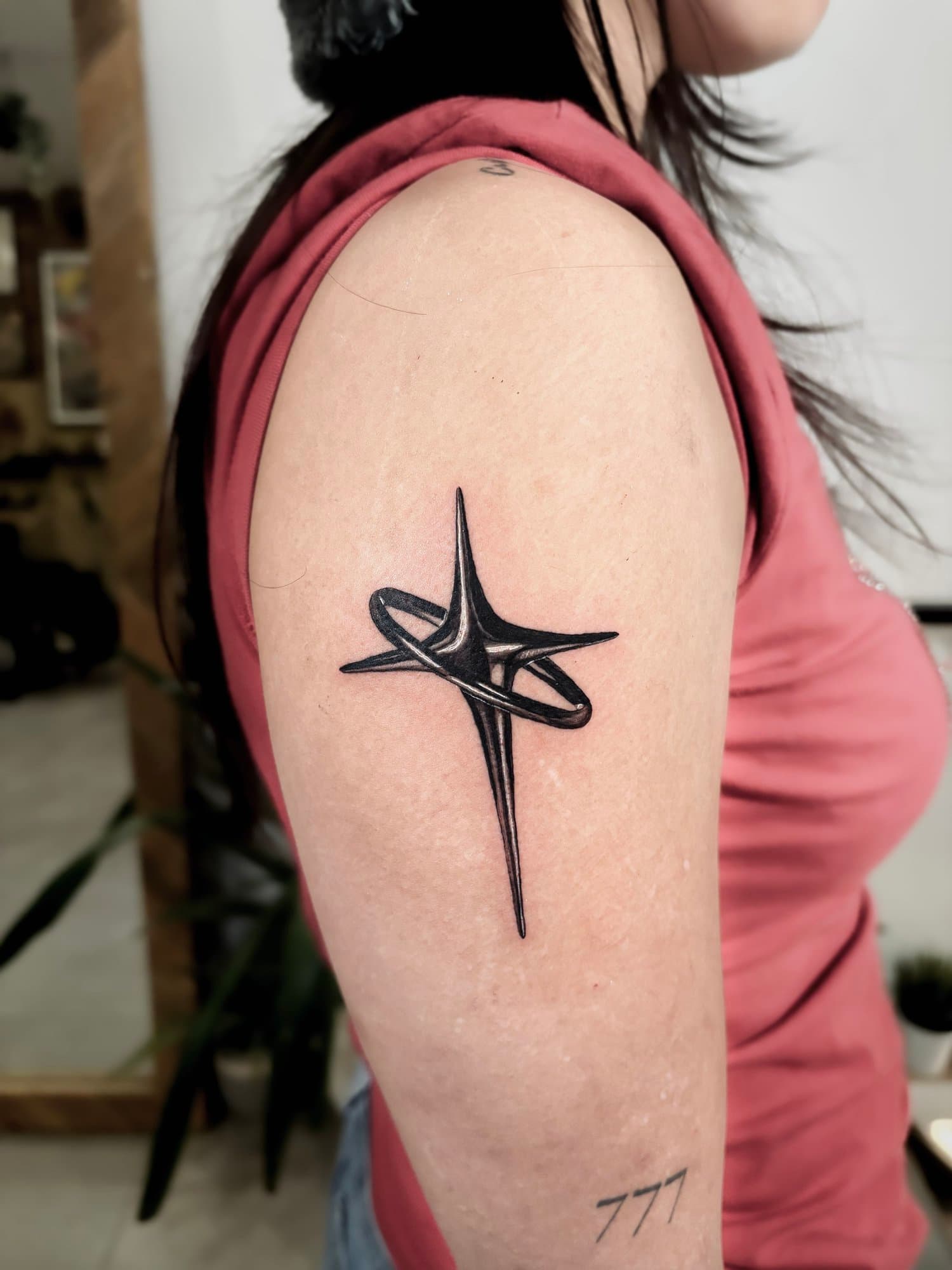 A close-up of a person's upper arm with a black star tattoo and script tattoo. The person is wearing a pink sleeveless top and is in an indoor setting. Part of