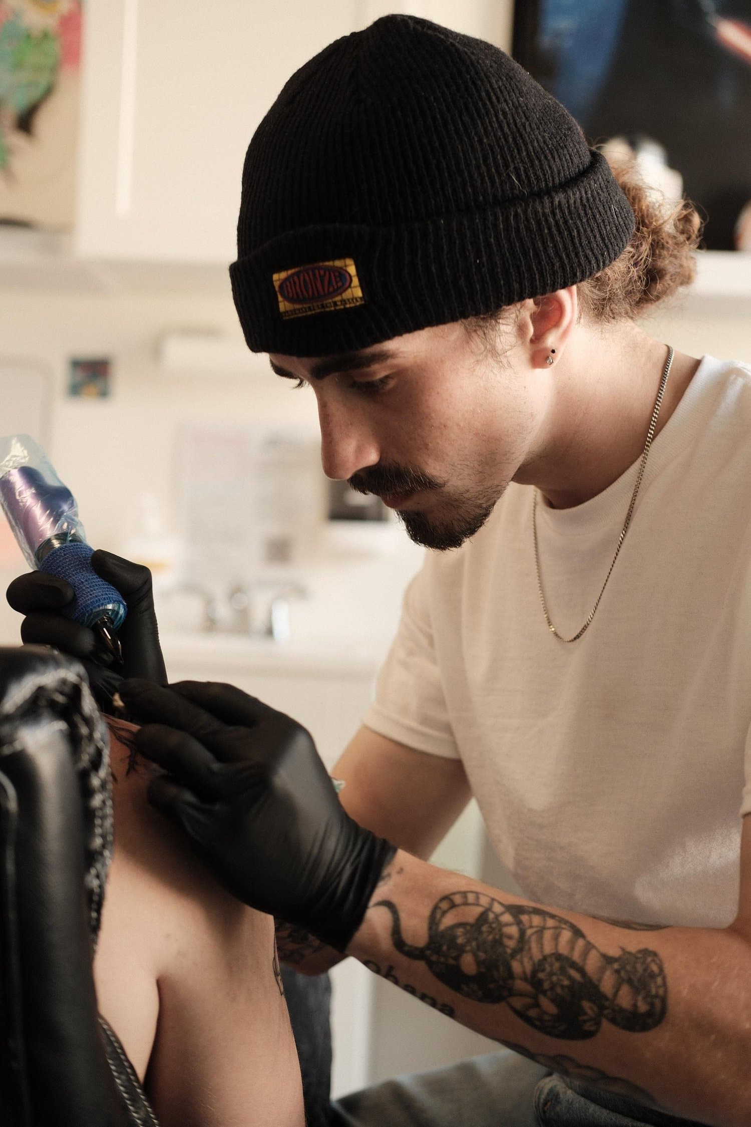 A man with a beard, wearing a black beanie, tattoos on his arm, and black gloves, tattooing a person's skin.