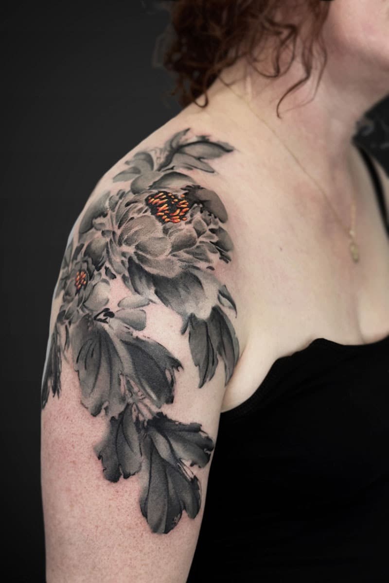 Grayscale floral tattoo on a woman's upper arm and shoulder, featuring large peony blossoms with small orange centers.