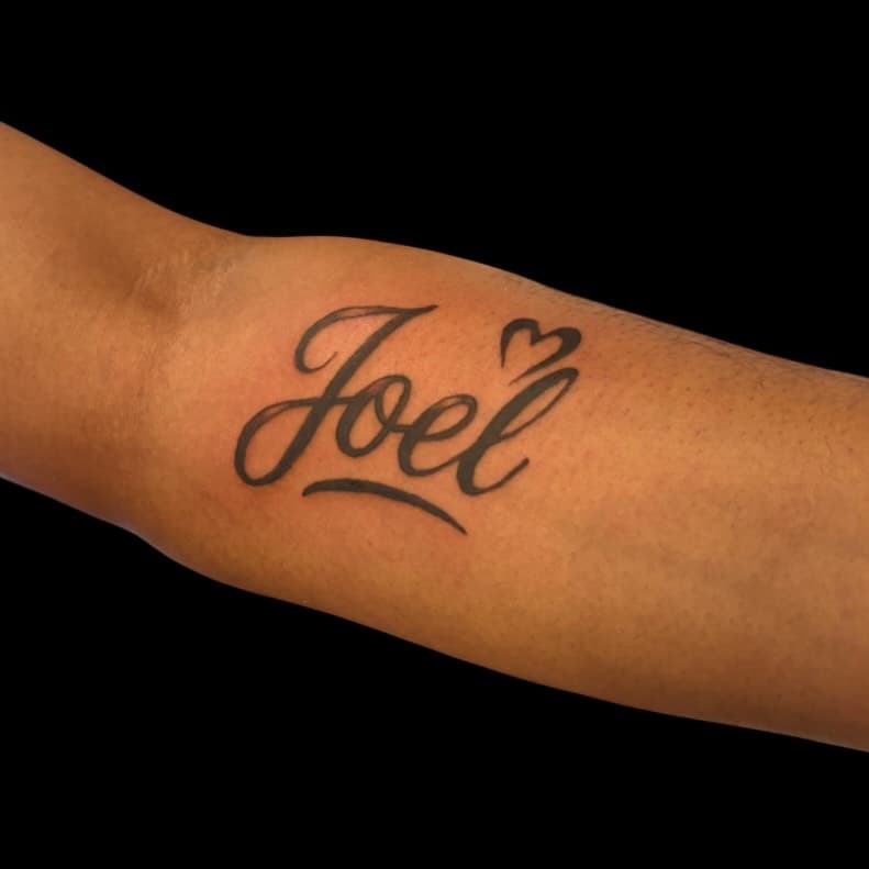 Close-up of a black ink tattoo of the name "Joel" with a small heart on a person's arm.