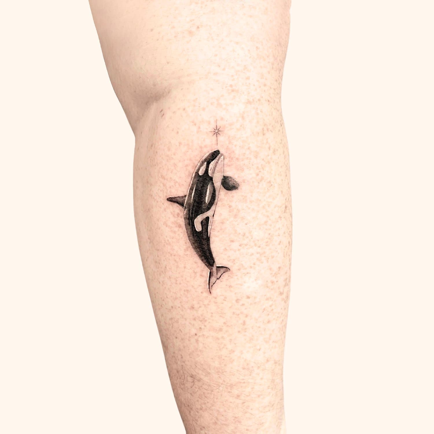 Tattoo of a orca whale swimming upward on person's thigh with a small black dot and a star above