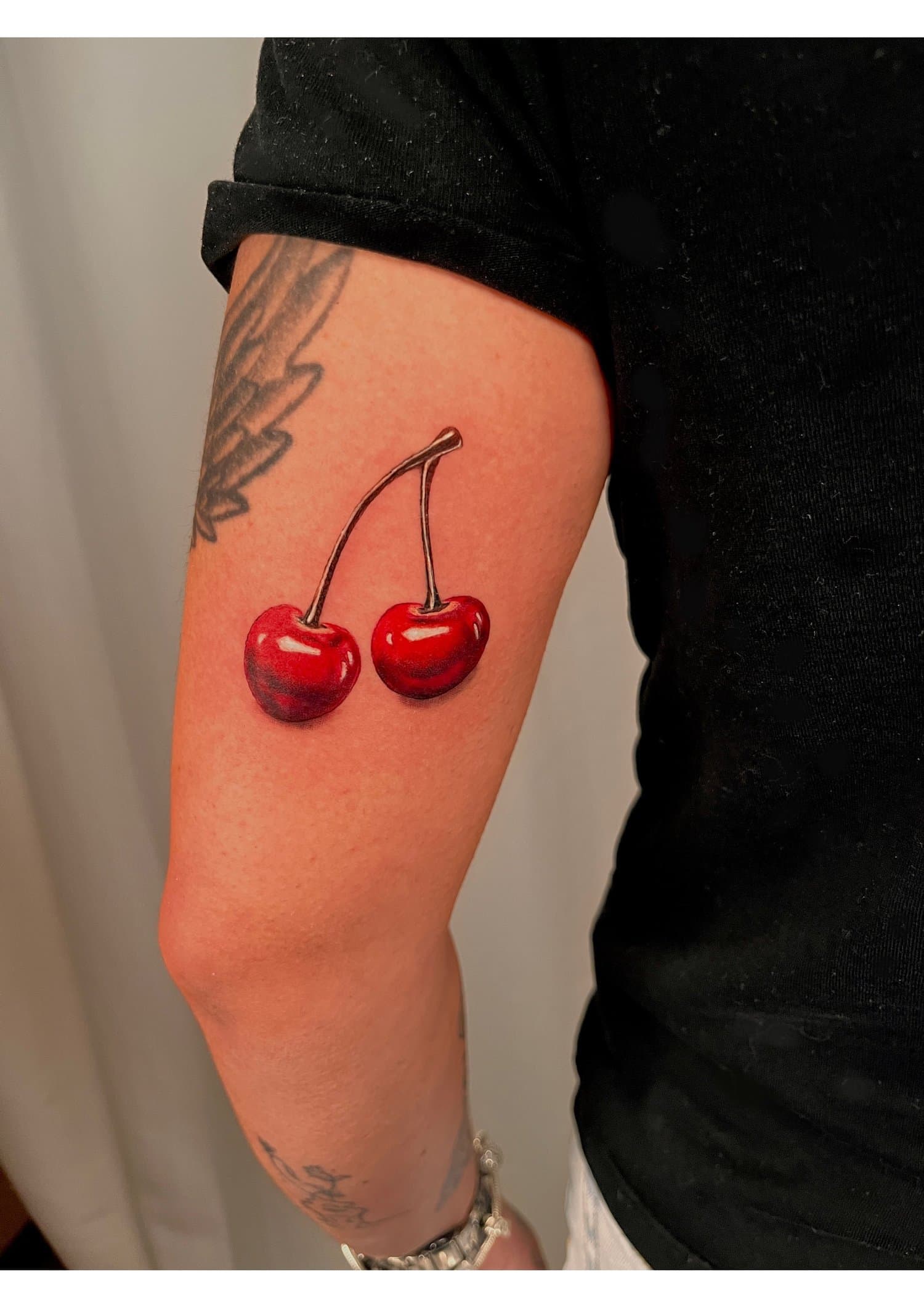 A person wearing a black rolled-up sleeve shirt, showing tattoos on their arm with cherries tattooed on the forearm, and jewelry including a bracelet and rings,