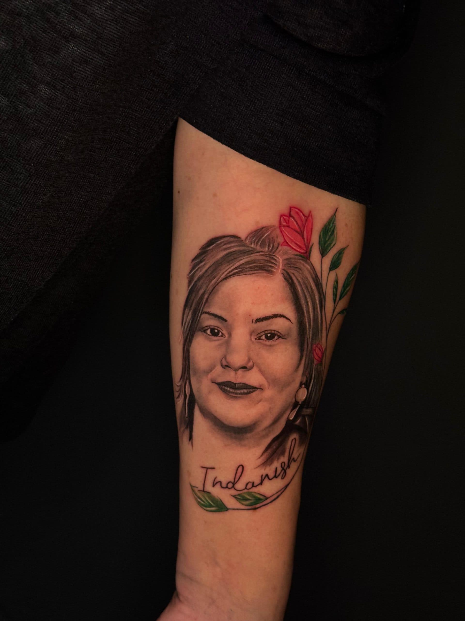 Portrait tattoo of a woman's face with red flowers and the name 'Indanish' in script on an arm.