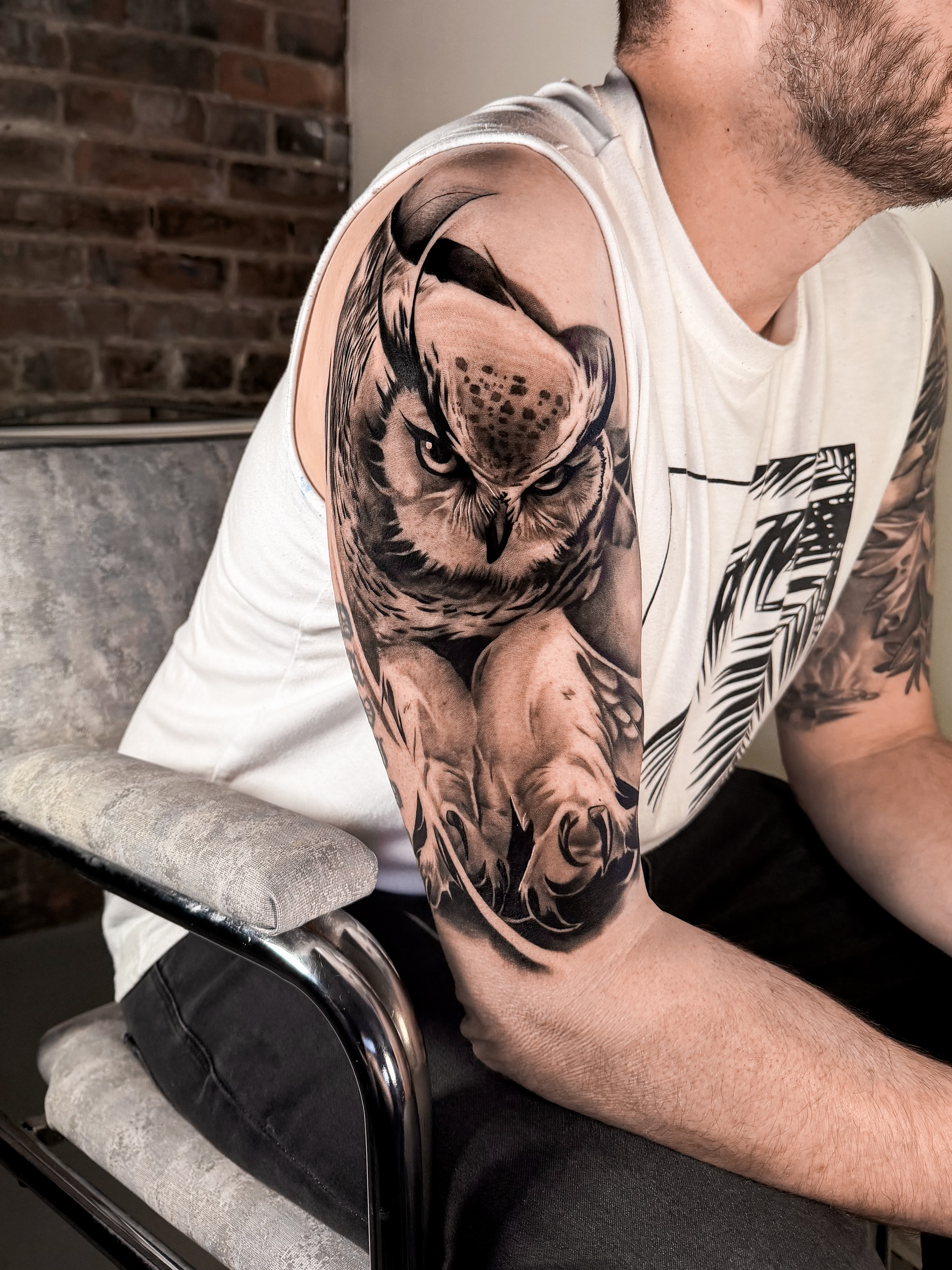 Large black-and-grey upper-arm tattoo of a forward-facing owl with extended talons