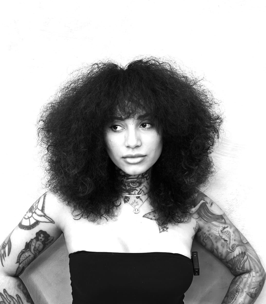 Zhaddi - Tattoo Artist - Black and white photo of a woman with voluminous curly hair, tattoos on her arms and neck, wearing a strapless top and jewelry, standin