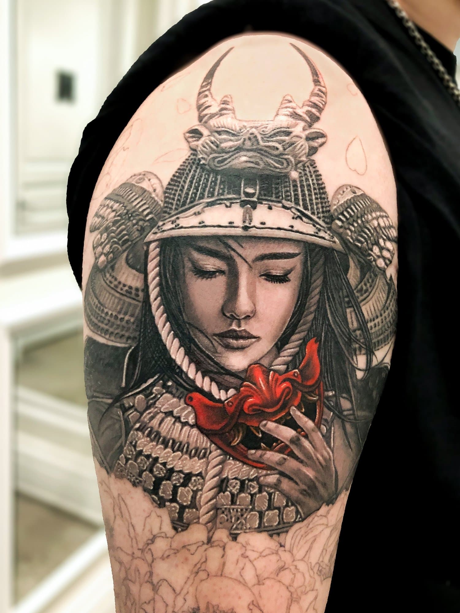 Tattoo of a woman with her eyes closed, wearing a samurai helmet with a fierce animal mask on top, adorned with armor and a red ribbon, on the upper arm.