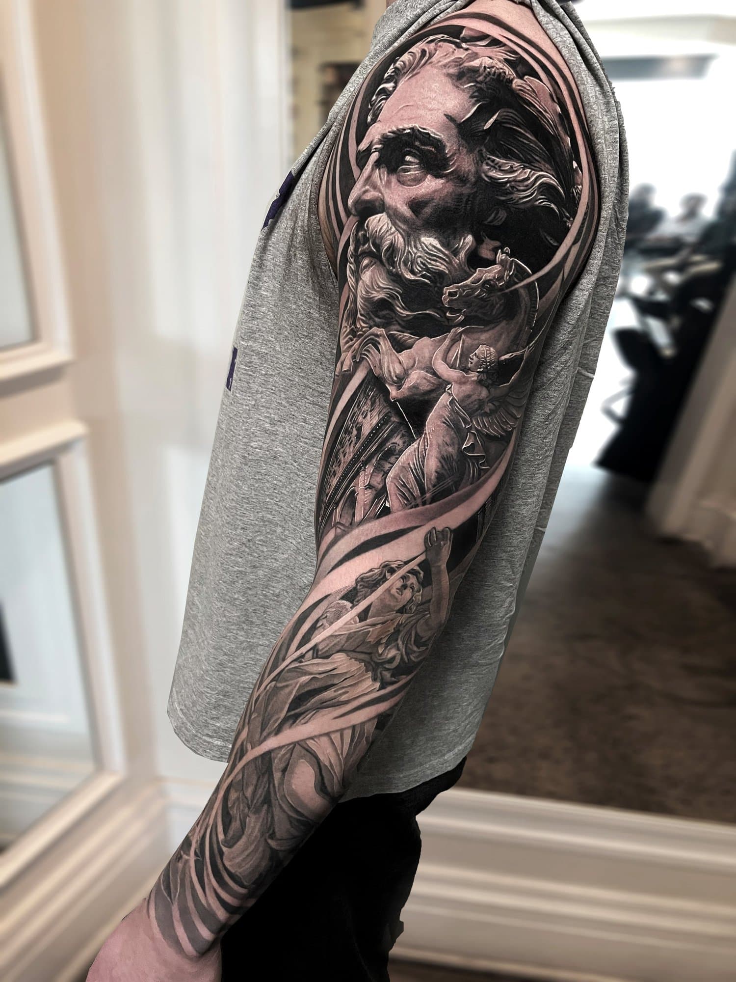 A person with a detailed black and gray tattoo sleeve featuring a face of a bearded man, a warrior with a spear, and dramatic shading on their arm.