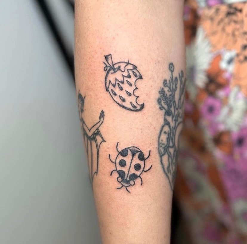 Tattoo of a ladybug, strawberry, and a partial drawing of a woman with a tree, on a person's arm.