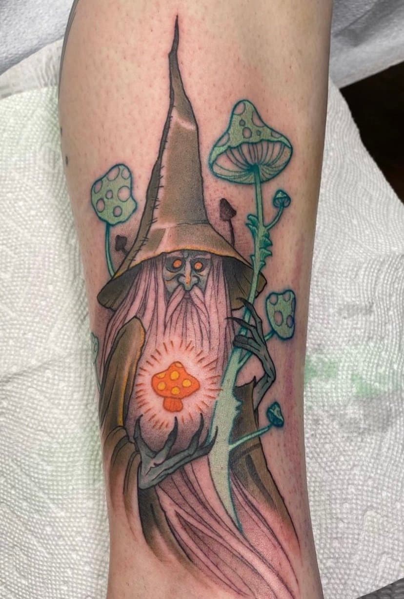 A tattoo of a wizard with a long beard, wearing a pointed hat, holding a glowing mushroom, surrounded by smaller mushrooms.