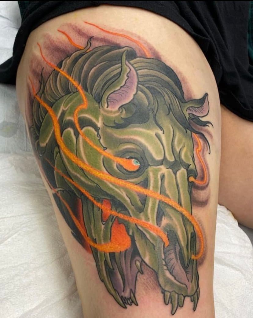Tattoo of a green dragon skull with orange accents on a person's thigh.