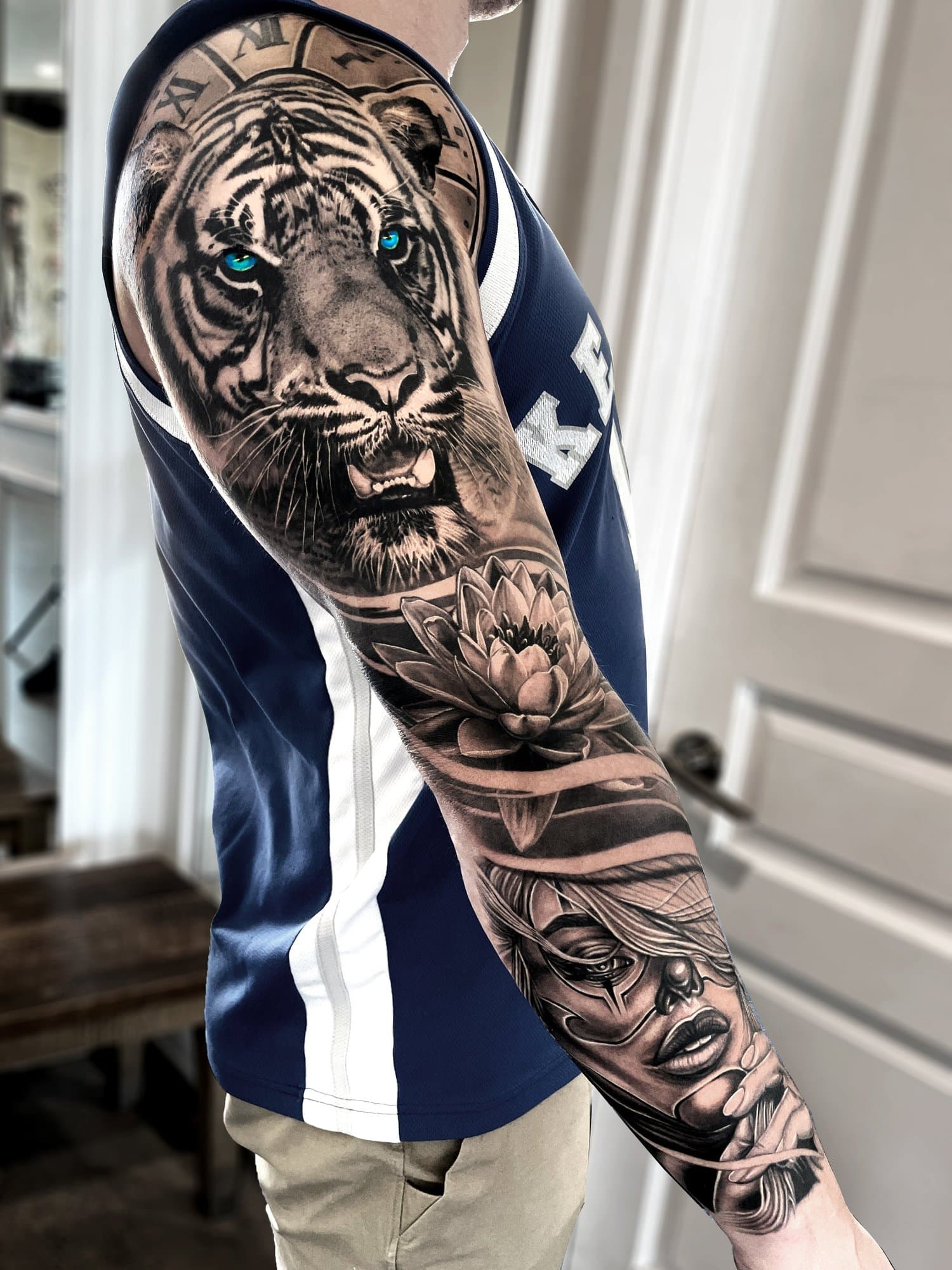 Black and grey tattoo sleeve featuring a tiger with piercing blue eyes, a lotus flower, and a woman's face with flowing hair.