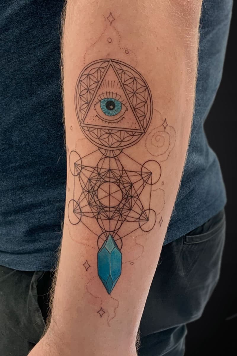 Geometric forearm tattoo with an eye inside a triangle within a circle, connected by Metatron's Cube lines and a blue crystal pendant at the bottom.