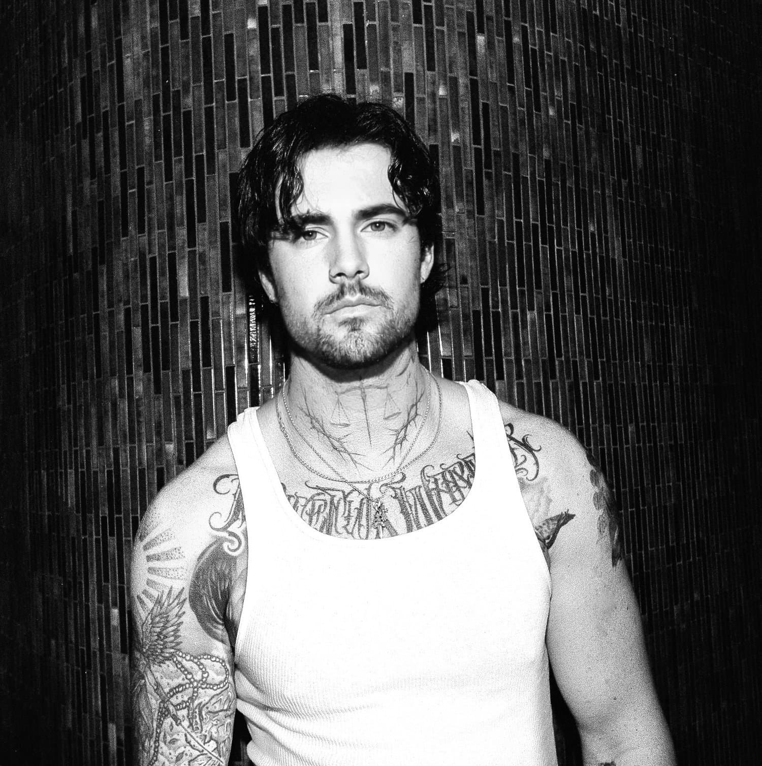 Mitch - Tattoo Artist - Black and white photo of a man with dark, wavy hair, a beard, and tattoos on his arms and chest, wearing a white tank top, standing in f