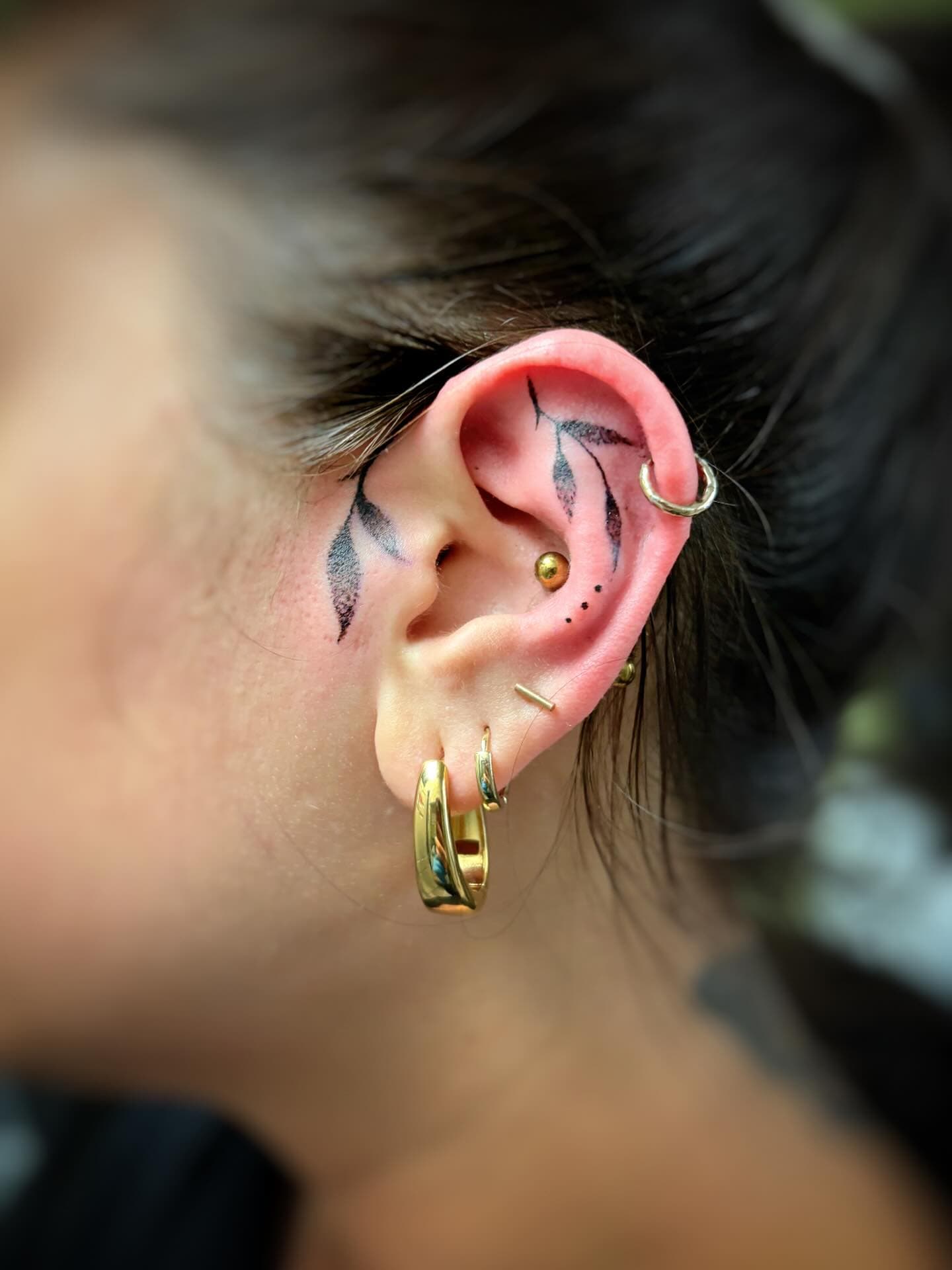 Close-up of a woman's ear with minimalist leaf tattoos and gold hoop earrings.