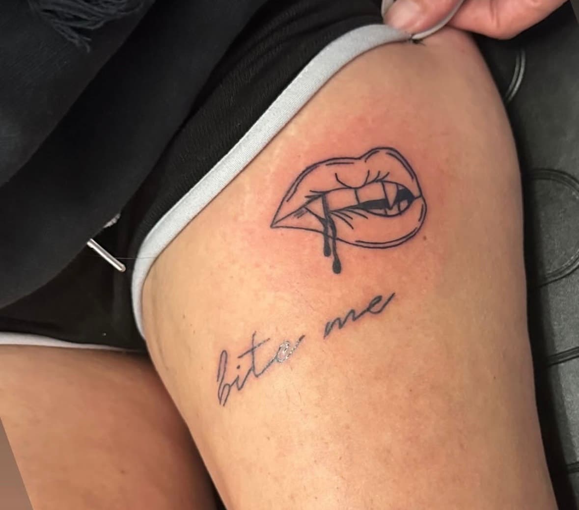 Fine line tattoo of teeth biting lip and script reading "bite me" beneath