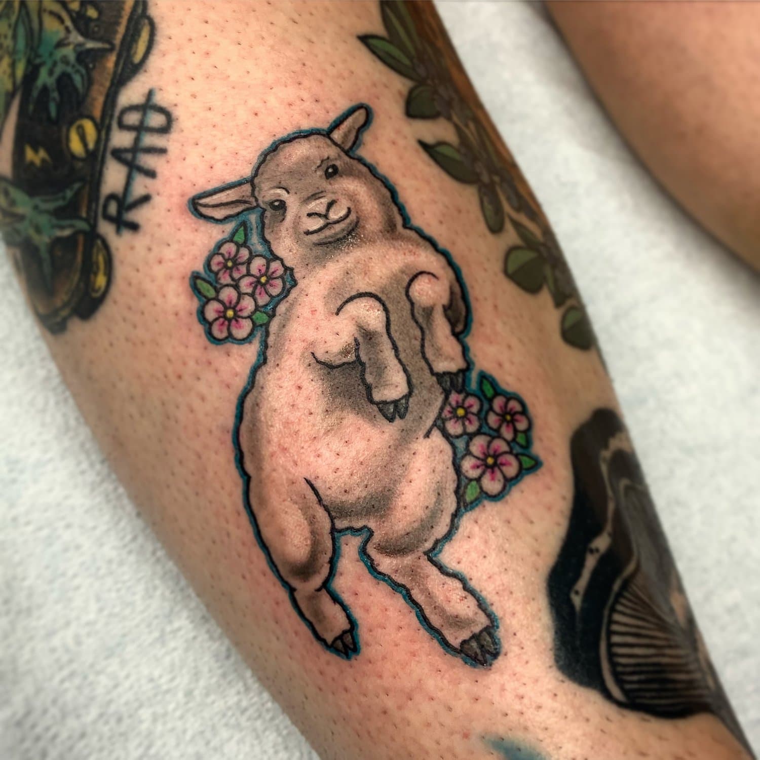 Tattoo of a smiling baby goat with pink flowers around it and a faint outline.